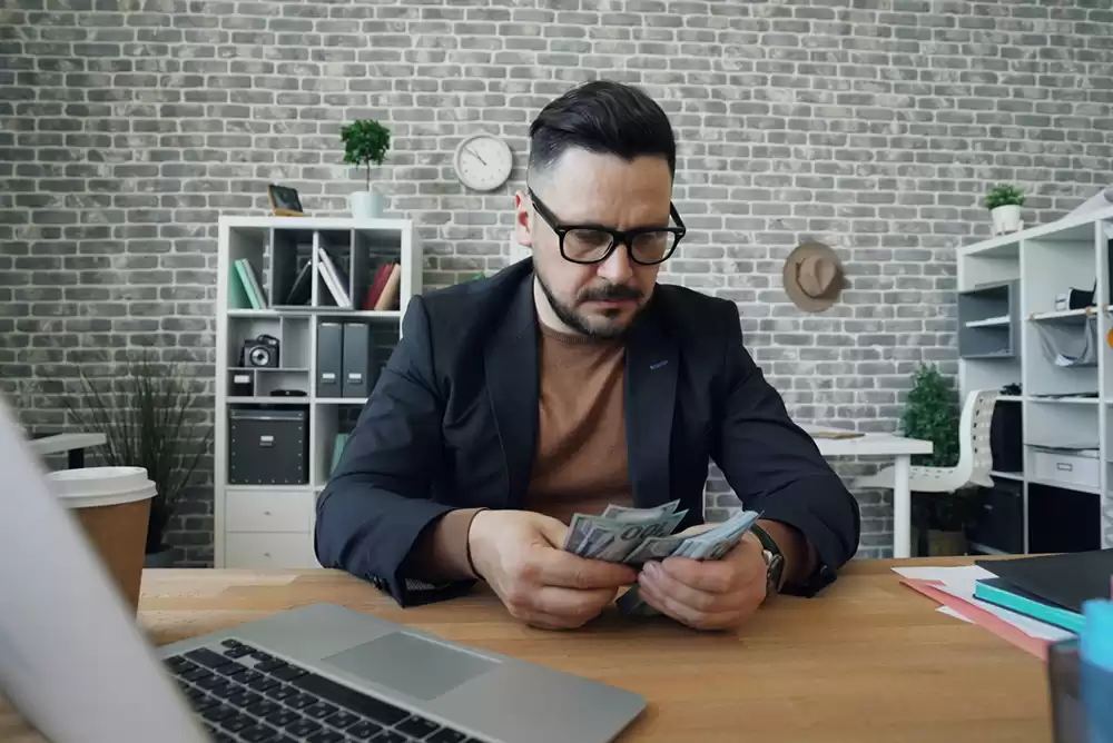 A man in a suit jacket sitting at a desk counting 100 dollar bills.