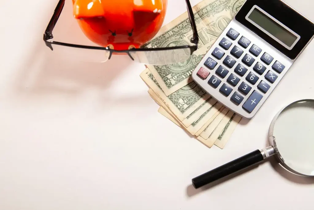 A piggy bank with reading glasses, dollar bills, a calculator, and a magnifying glass on a white surface.